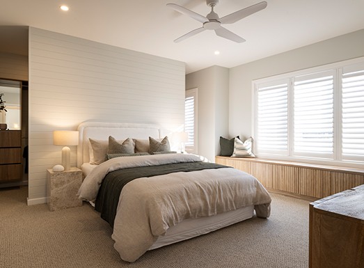 Modern bedroom with a white upholstered bed centered against a textured accent wall, dressed in beige and dark bedding with decorative pillows. Nightstands with warm table lamps flank the bed. Large white shuttered windows overlook the room. A ceiling fan hangs above, and neutral carpeting covers the floor. Dark wood furniture visible on left and right sides.
