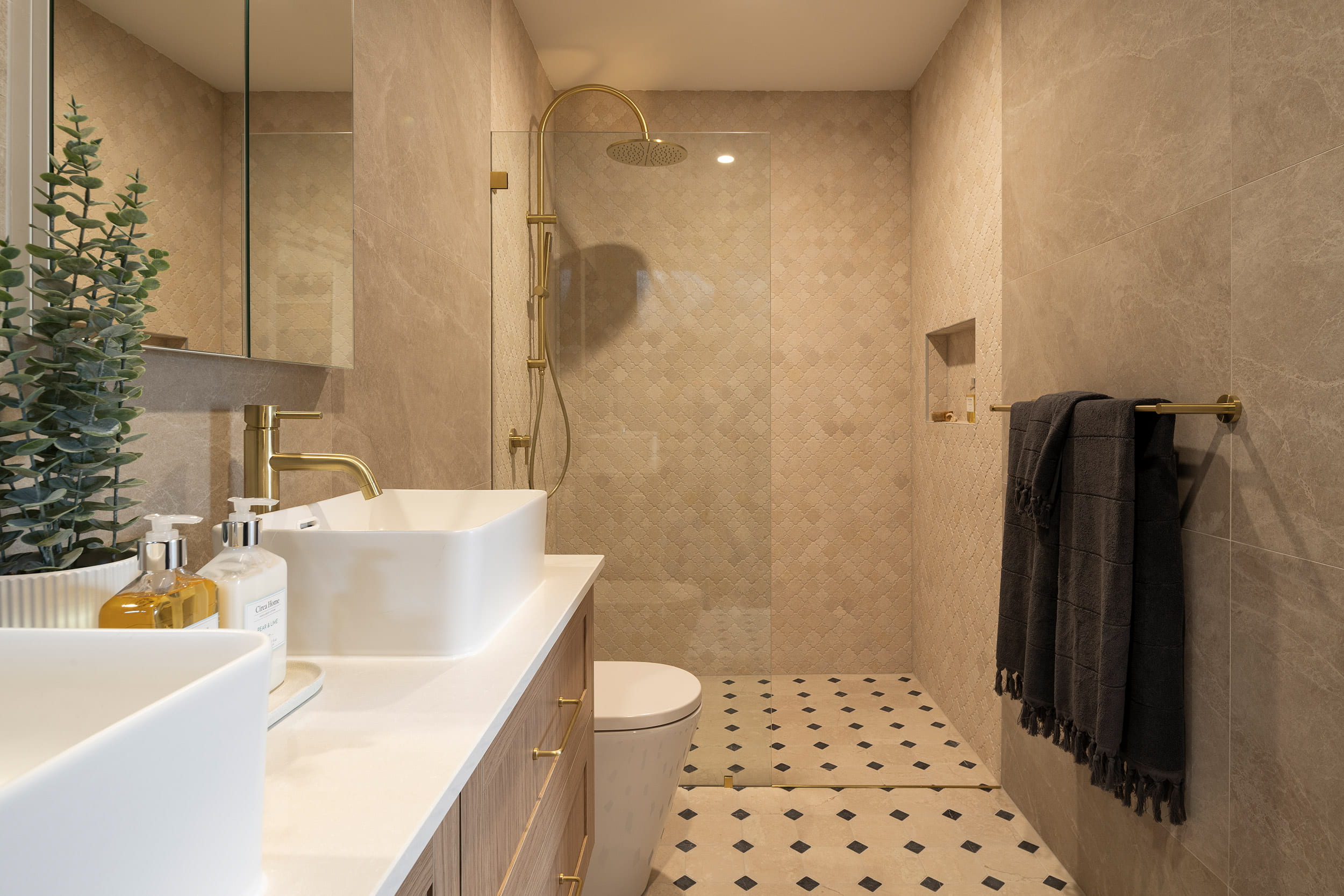 Modern ensuite bathroom featuring a side view of a spacious walk-in shower with brass fixtures and rainfall showerhead, beige tiled walls, white vanity with two vessel sinks, brass tap fixtures, decorative plant, and black and white diamond-patterned floor tiles. Dark towels hang on a brass rail on the right side.