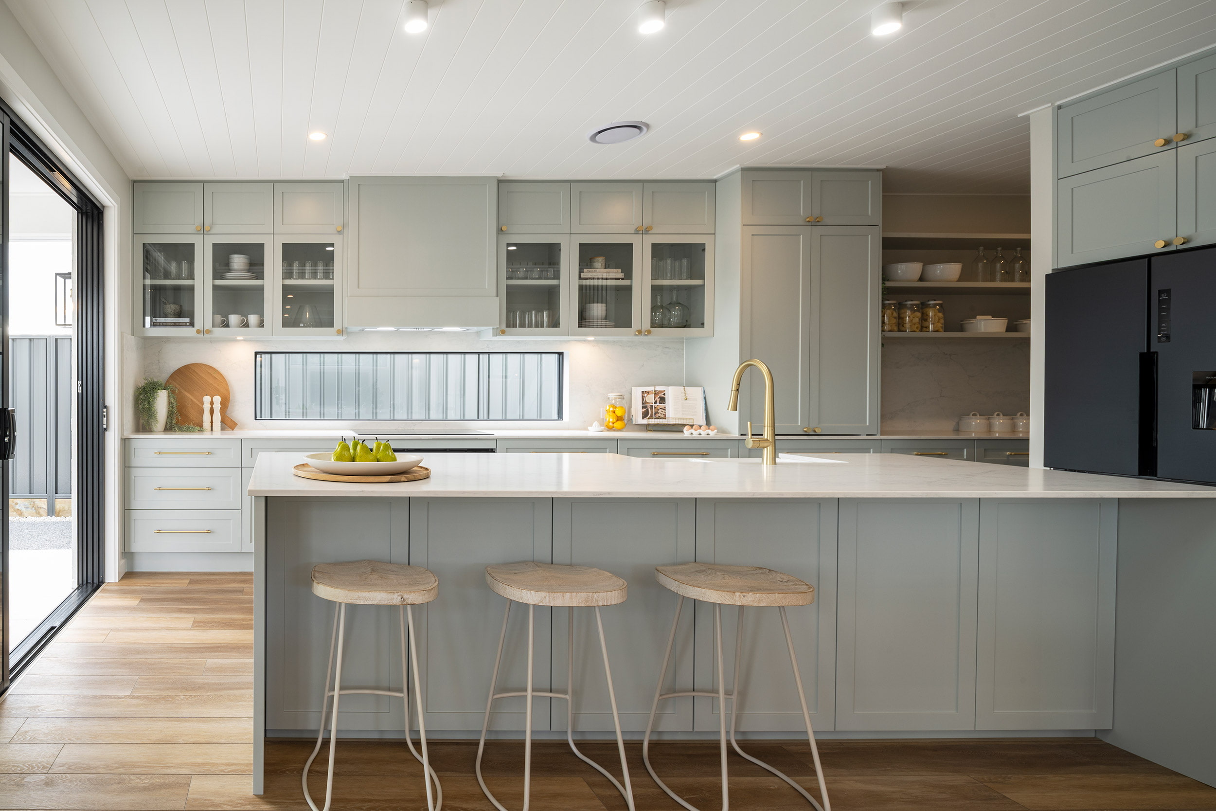 Modern kitchen with sage green and white cabinetry, white countertops, gold hardware, and a large island with three wooden bar stools. Features glass-front upper cabinets, a gold faucet, black appliances on the right, recessed lighting, and hardwood flooring. A sliding glass door opens to the left, and the kitchen displays decorative dishware and fresh pears on the counter.