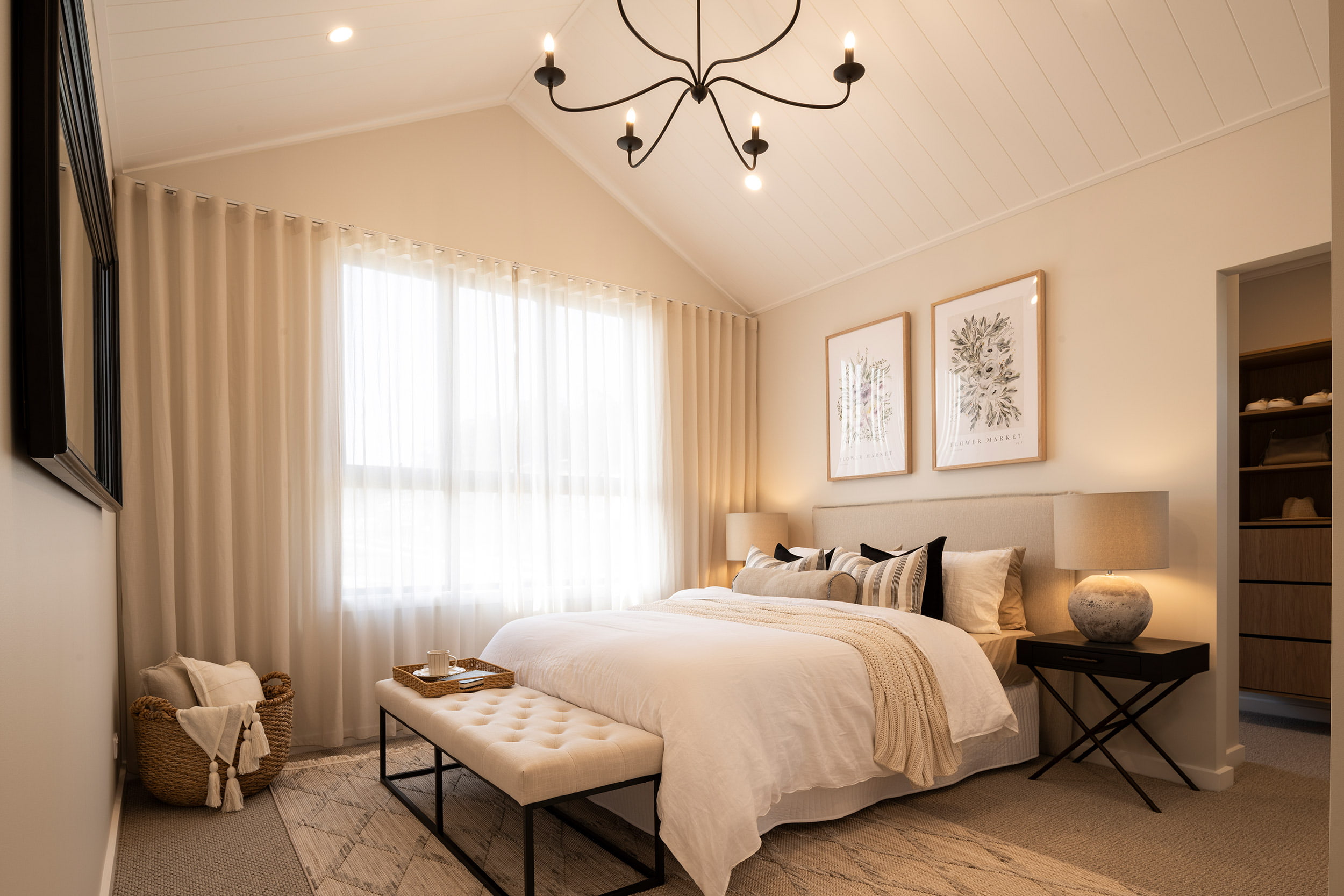 Master bedroom in Montgomery Homes Waverley display home featuring a bed with neutral bedding and decorative pillows, upholstered headboard, black wrought iron chandelier, cream curtains over large windows, botanical wall art, bedside tables with lamps, upholstered bench at foot of bed, and neutral color palette throughout.