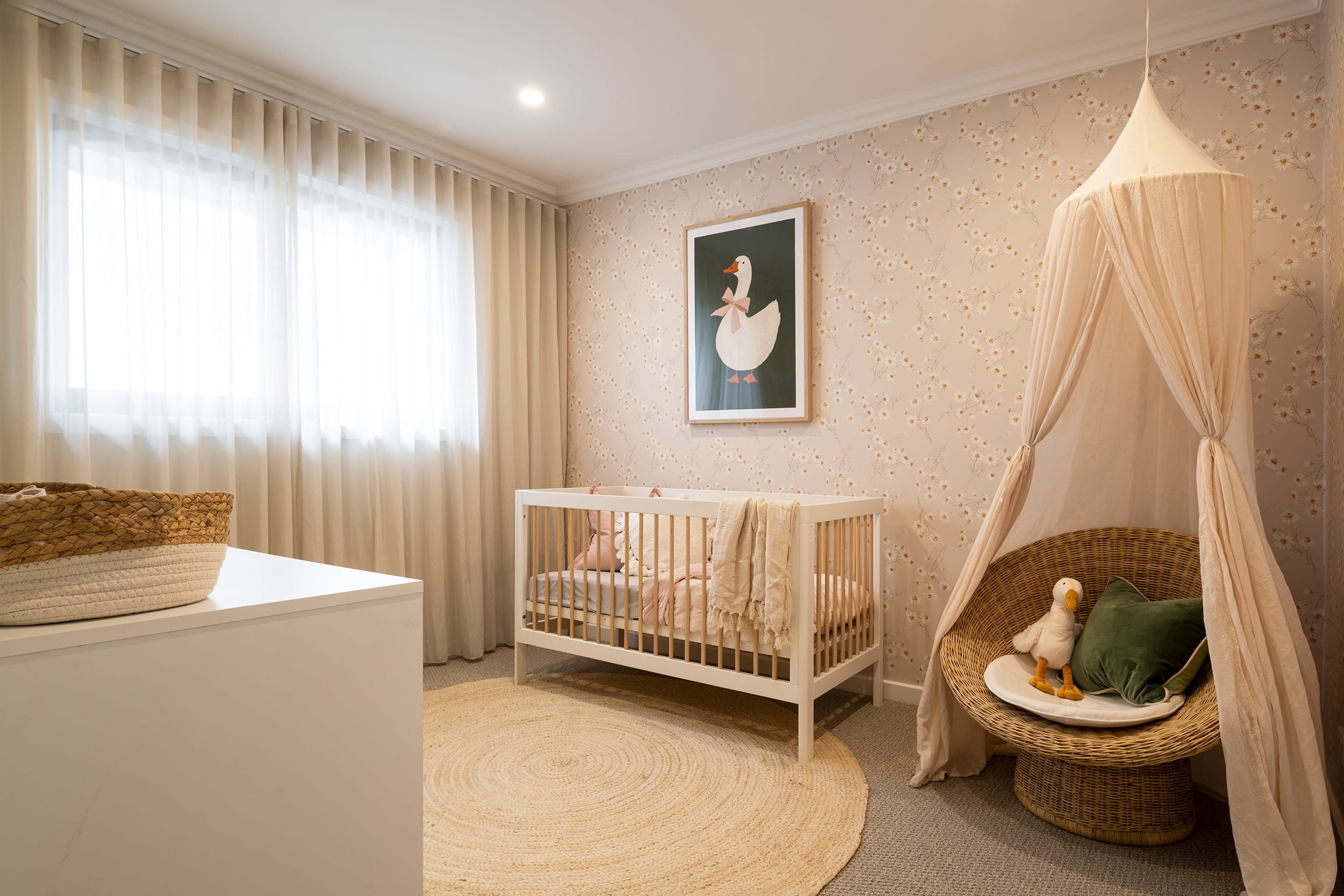 Modern nursery room with neutral beige and cream tones, featuring a white wooden crib with bedding, a woven rattan canopy chair with cushions, patterned wallpaper with small dots, floor-to-ceiling curtains, a round woven rug, framed duck artwork on the wall, and natural lighting from large windows.