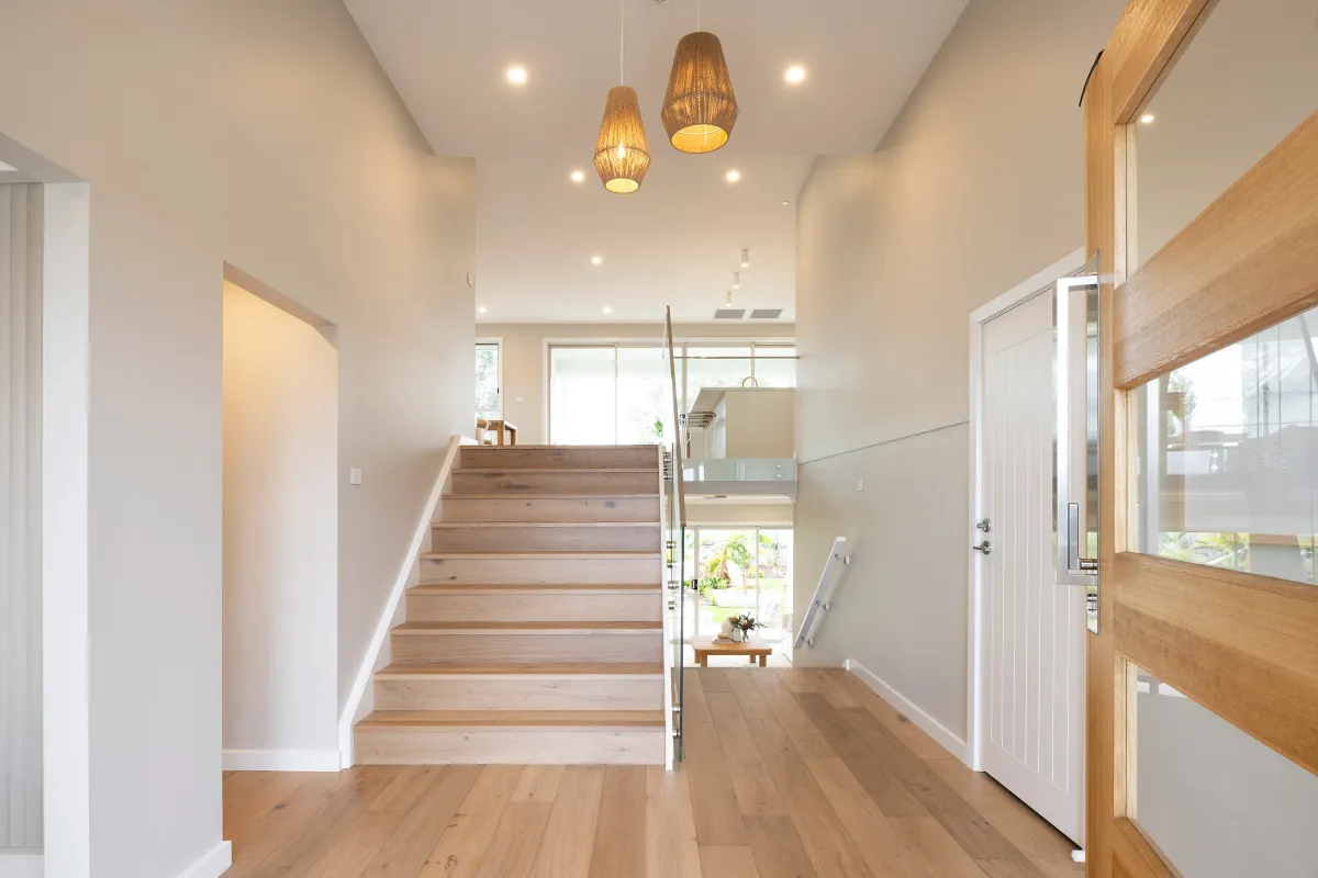 Modern home entryway featuring a wooden staircase with glass railing, light wood flooring, cream-colored walls, and two woven pendant lights hanging from the ceiling. Natural light streams through large windows and glass doors leading to an outdoor space.
