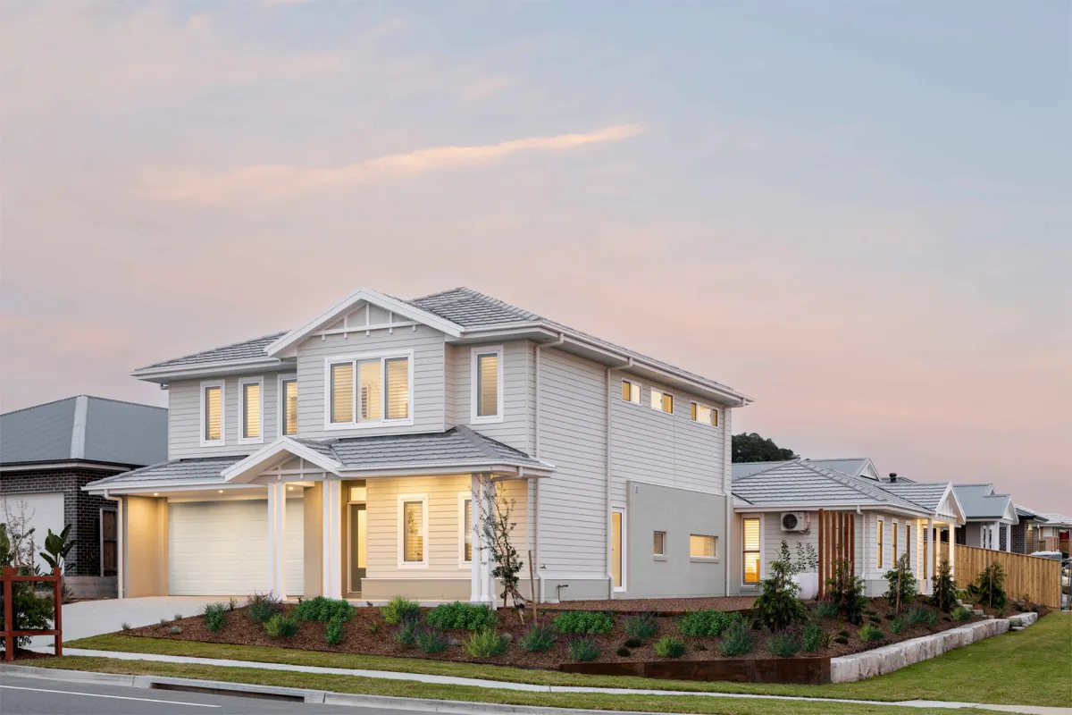 Modern two-story white residential home at dusk with illuminated windows, featuring a double garage, pitched roof with grey tiles, landscaped front garden with young plants and shrubs, and neighboring houses visible to the right, taken in Newport.