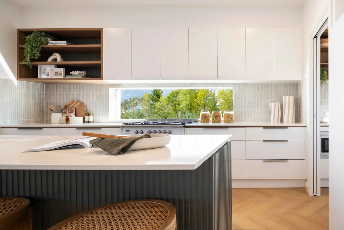 Modern kitchen with white cabinetry and wood accents, featuring a large window overlooking green trees, a gas cooktop, wooden open shelving with decorative items, a white island with dark ribbed base, white subway tile backsplash, and wooden flooring.
