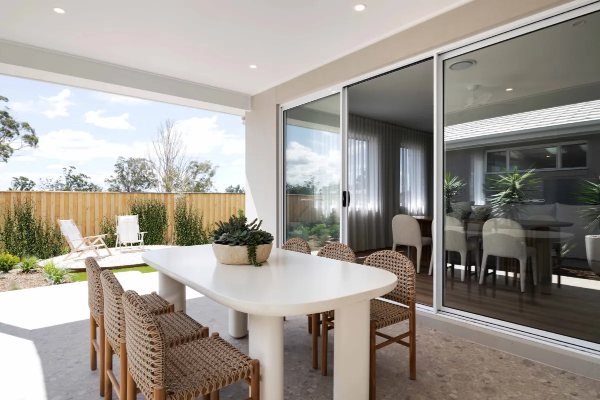 Modern outdoor entertaining space with a white oval dining table surrounded by woven chairs, large sliding glass doors opening to an interior living area, and a landscaped backyard featuring wooden fencing, lounge chairs, and planted gardens under a cloudy sky.