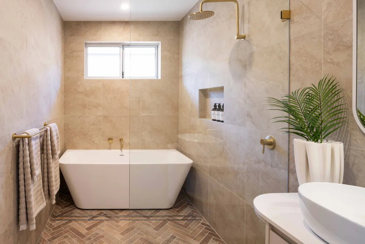 Modern bathroom with a white freestanding soaking tub centered in the room, surrounded by beige travertine tile walls. Brass fixtures and showerhead visible above the tub. A walk-in shower with glass enclosure is on the right side. Herringbone pattern tile flooring in brown tones. Small window above the tub, built-in niche with toiletries, potted palm plant, and towel rack with rolled towels on the
