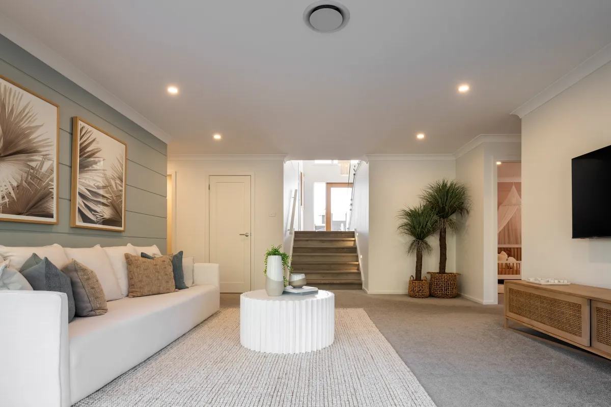 Modern living room with sage green shiplap accent wall featuring framed botanical artwork, white upholstered sofa with decorative pillows, round white ribbed coffee table, potted palm plant, wooden media console with rattan doors, recessed lighting, and wooden staircase leading to upper level.