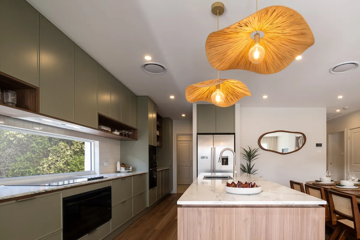 Modern kitchen with olive green cabinetry, white marble countertops, and a central island. Two organic-shaped wooden pendant lights with warm bulbs hang above. A window with garden view overlooks the sink area on the left. Stainless steel appliances, a decorative bowl with dried flowers, and wooden dining chairs with cane backs are visible on the right. Recessed lighting and a bronze-framed mirror accent the space.