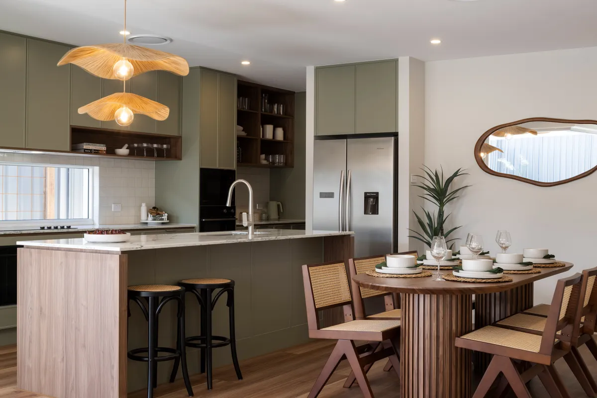 Modern kitchen and dining area featuring sage green cabinetry with open shelving, two sculptural wing-shaped pendant lights, stainless steel refrigerator, marble countertops, and a kitchen island with bar seating. A wooden dining table with pink chairs and place settings sits adjacent to the kitchen, with a decorative oval mirror and potted plant on the wall.