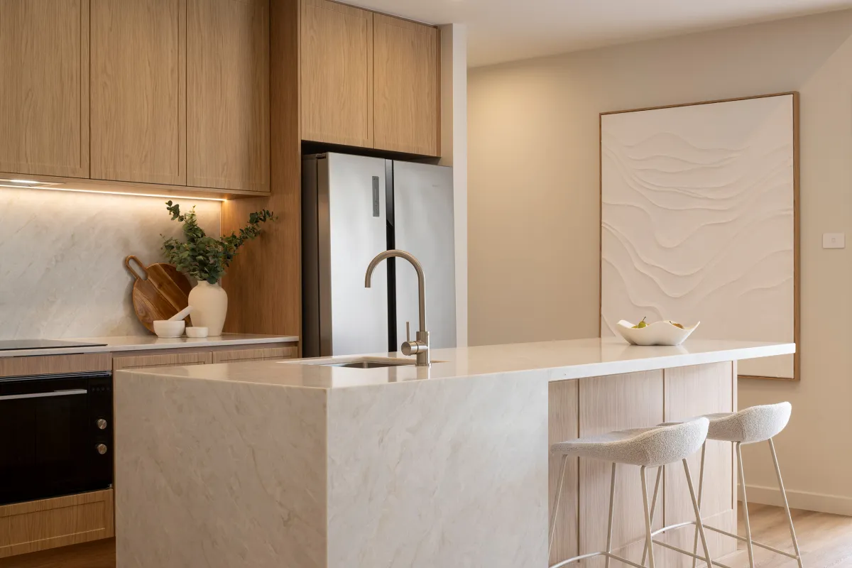 Modern kitchen with light oak cabinetry, white marble island with waterfall edge and integrated sink, stainless steel faucet, two beige upholstered bar stools, stainless steel refrigerator, and decorative textured wall art above the island seating area.