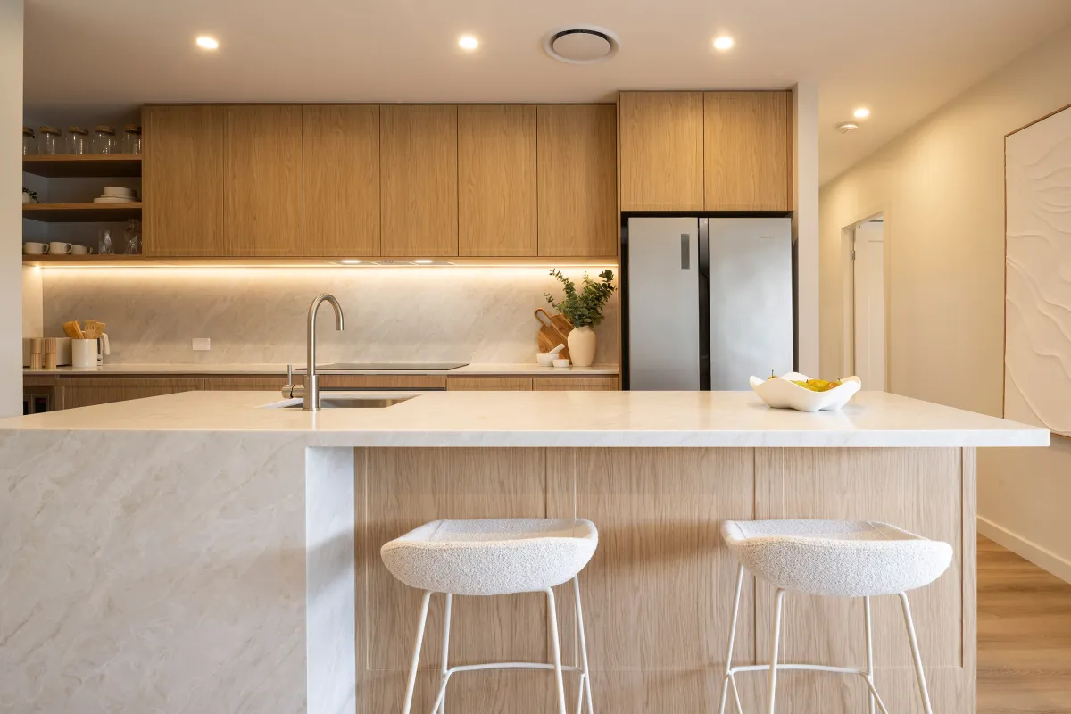 Modern kitchen with light wood cabinetry, white marble countertops and backsplash, stainless steel sink and faucet, black and stainless steel refrigerator, recessed lighting, open shelving with dishware, and two cream-colored upholstered bar stools at the island.