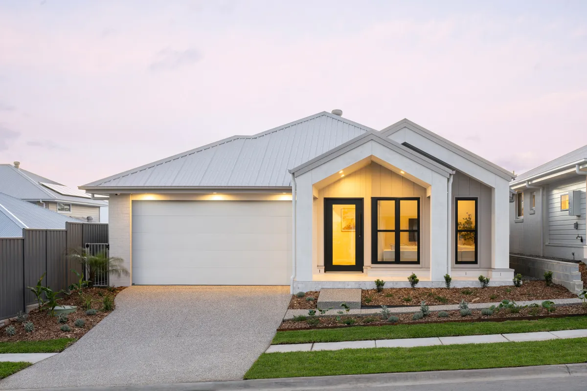 Modern white single-story house with metal roof, black-framed windows and doors with warm interior lighting, attached garage, concrete driveway, landscaped front yard with mulch and young plants, neighboring houses visible, twilight sky.