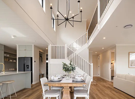 3D virtual tour of a modern Newcastle home interior featuring an open-plan living space with a two-story staircase, dining table with white chairs, contemporary kitchen with stainless steel appliances, wooden flooring, and a geometric pendant light fixture hanging from the ceiling.