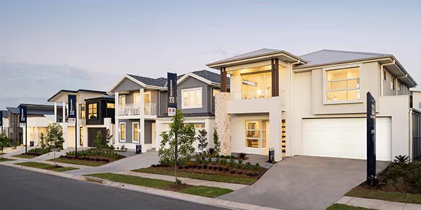 A row of modern display homes with contemporary architecture featuring cream and dark facades, illuminated windows at dusk, manicured lawns, driveways, and black accent pillars along a residential street.