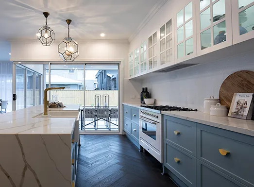 Modern kitchen interior with blue-grey cabinetry, marble countertops, stainless steel appliances, geometric pendant lights, and floor-to-ceiling windows overlooking Sydney cityscape.