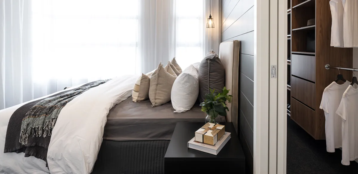 Modern bedroom with a black bed dressed in white linens and multiple neutral-toned pillows, a dark textured throw blanket, and a woven accent piece. Large windows with sheer white curtains provide natural light. A black nightstand holds decorative gold boxes. A pendant light hangs above the headboard. To the right, a dark wood walk-in closet with open shelving displays white clothing and folded items. A small potted plant sits on the night