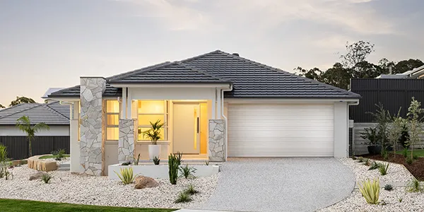 Modern single-story display home with white and dark grey facade, featuring a curved roofline, large front windows with warm lighting, white garage door, stone pillars flanking the entry, and landscaped front yard with gravel and native plants in Warnervale.