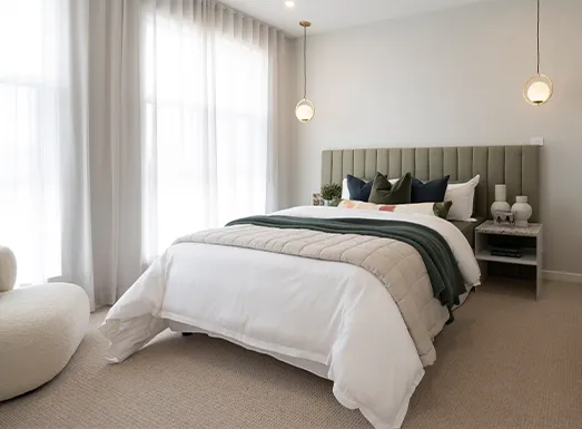 Modern bedroom with a gray upholstered headboard bed dressed in white and green bedding, pendant lights overhead, sheer curtains covering large windows, a round white ottoman in the foreground, nightstand with decor, and neutral beige carpet flooring.