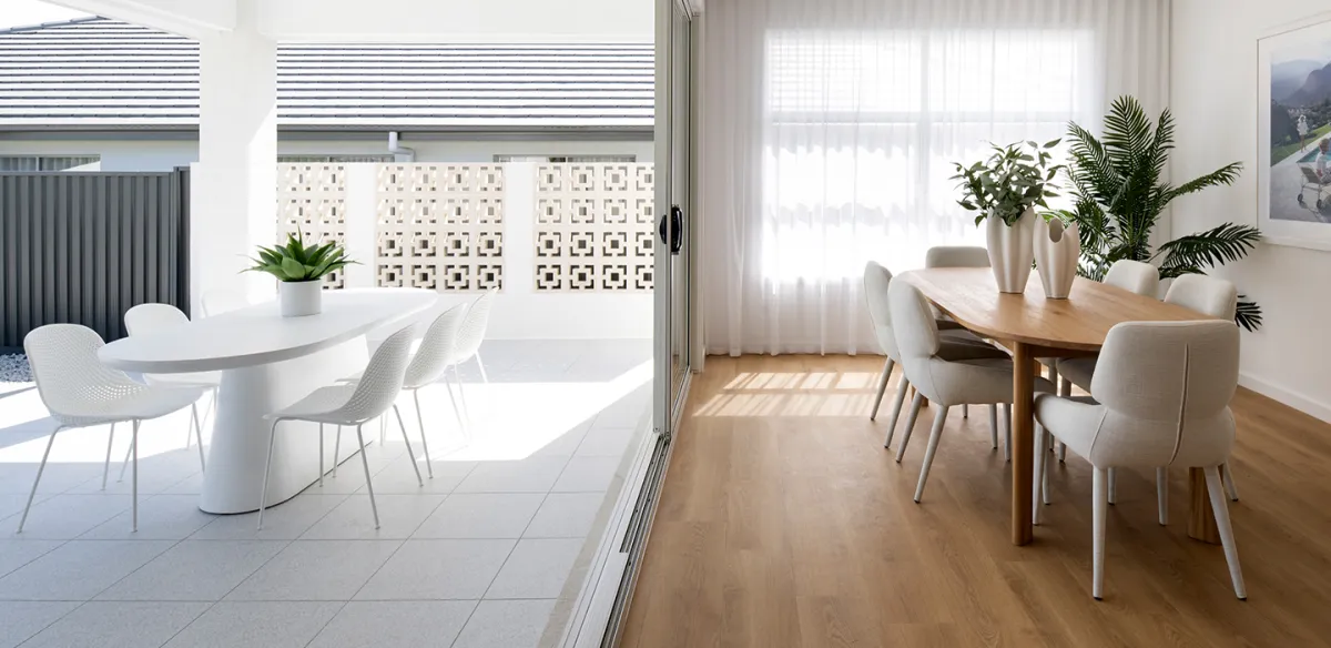 Modern dining spaces at Avoca Warnervale featuring two distinct areas: left side with white round table and chairs on tiled flooring opening to a balcony with geometric screen; right side with wooden dining table, cream upholstered chairs, potted plants, and wooden flooring with natural light from large windows.