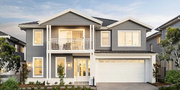 Modern two-story residential home with gray and white exterior, featuring a front-facing garage, upper-level balcony, multiple windows with warm lighting, and landscaped front yard with shrubs and trees.