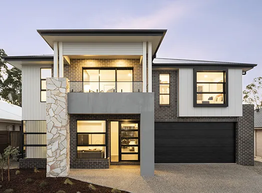 Modern two-story residential house with white and dark brick facade, large black-framed windows, a prominent stone accent column, black garage door, and illuminated interior spaces at dusk.