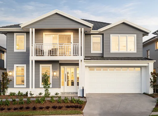 Modern two-story residential home with gray horizontal siding, white trim, and white railings. Features a two-car garage, front balcony, multiple large windows with warm interior lighting, and a concrete driveway. Landscaping includes small shrubs along the foundation and young trees on either side of the entrance.