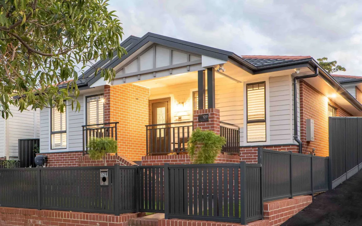 Modern residential home with contemporary heritage design featuring red brick base and white weatherboard upper sections, dark metal roof, large windows with shutters, covered front porch with dark metal railings, and black metal fence enclosure. Address number 129 visible on brick pillar. Green foliage visible on left side.