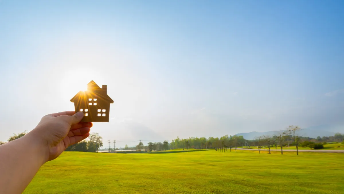 Hand holding wooden house model against sunlight in green field, symbolizing eco-friendly housing