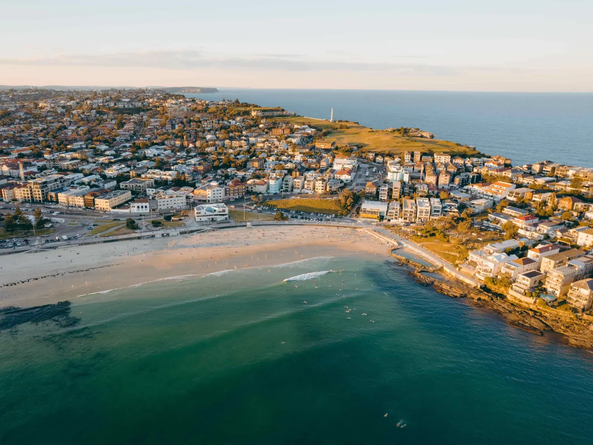 Aerial view of Bondi Beach at sunset, showing the iconic crescent-shaped sandy beach with turquoise water, surrounded by residential buildings and cliffs. The beachfront is lined with apartments and houses, with a headland lighthouse visible in the distance, and the Sydney coastline extending into the background under a clear sky.