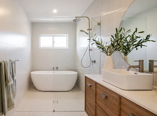 Modern bathroom featuring a white freestanding soaking tub centered in the space, a walk-in shower with glass enclosure and brushed gold fixtures, a wooden vanity with white vessel sink on the right, potted greenery in a cream vase, recessed lighting, and neutral tile flooring with a small window above the tub.