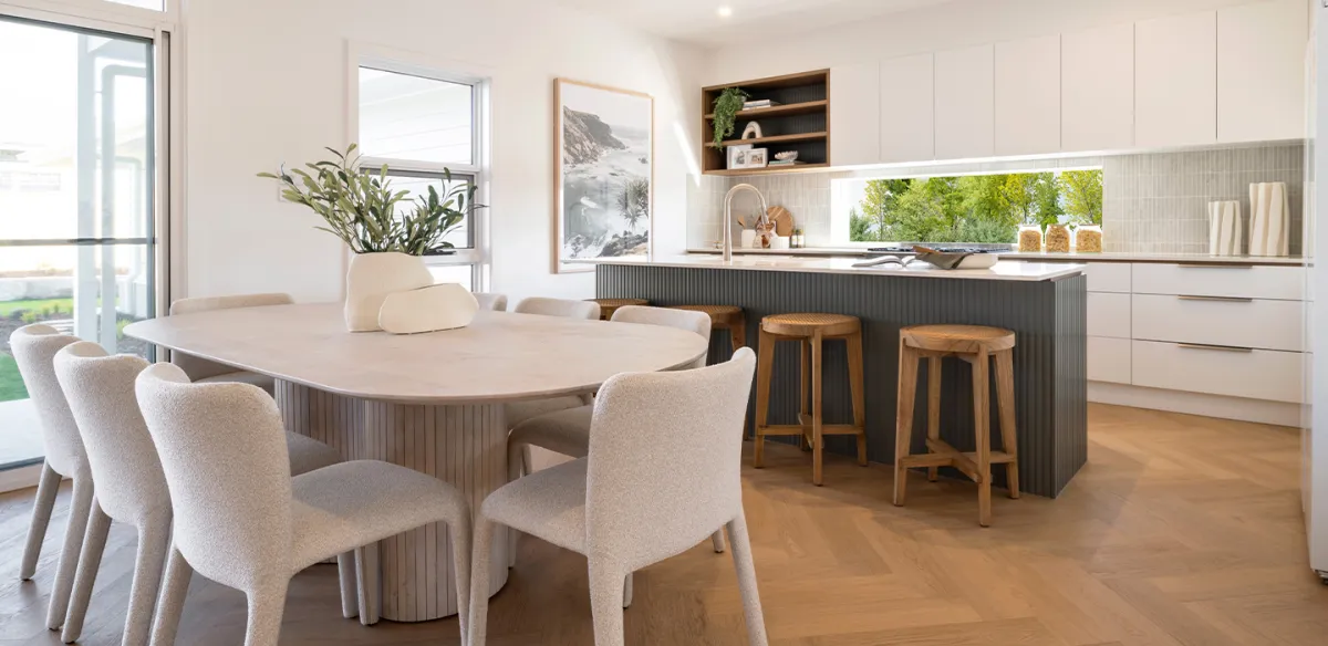 Modern open-plan living and kitchen space featuring a curved cream dining table with upholstered beige chairs, a dark kitchen island with wooden bar stools, white cabinetry, and large windows overlooking green gardens. Decor includes potted plants, framed coastal artwork, and wooden shelving.