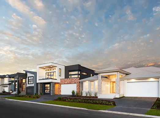 Modern residential building facade at dusk featuring contemporary architecture with mixed materials including white render, black cladding, and exposed brick accents. The multi-level home has large windows with warm interior lighting, a dark driveway, manicured landscaping, and is photographed against a dramatic cloudy sky at sunset.