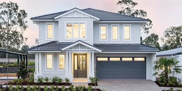 Modern two-story display home with white exterior, dark roof, and dark gray garage door. Large windows illuminate the facade at dusk. Landscaped front yard with plants and shrubs flanks the paved driveway. Gated fence visible to the left.