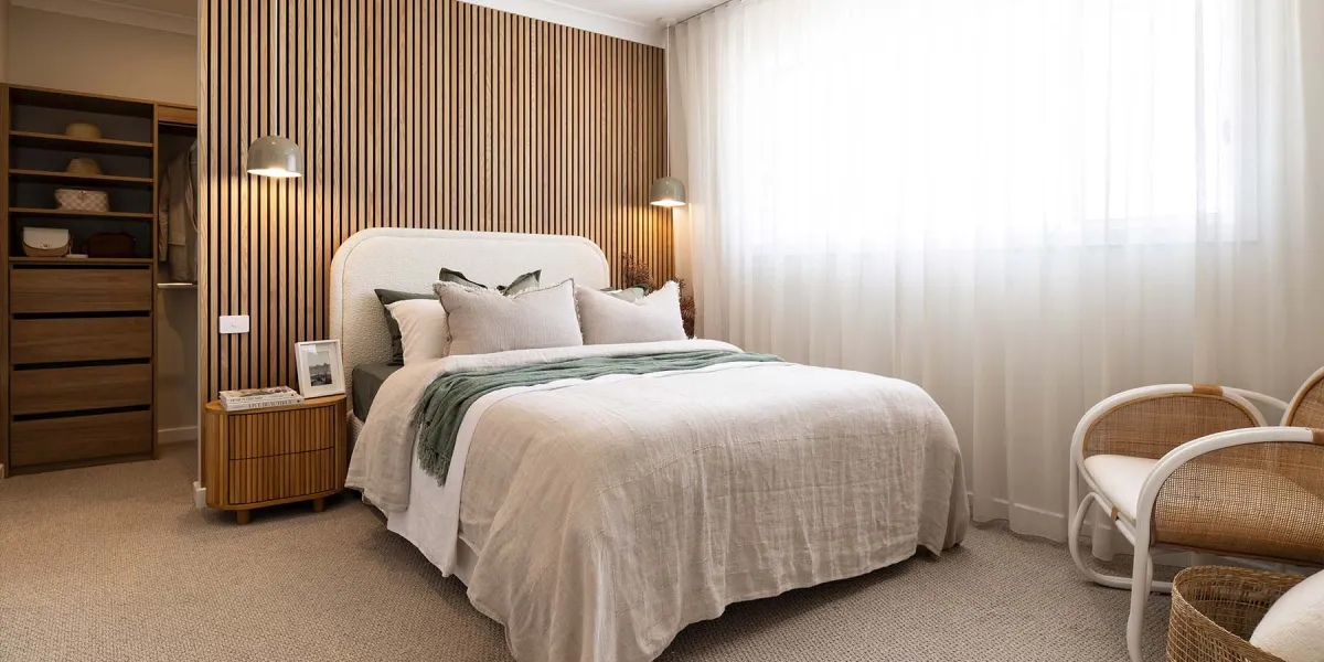 Master bedroom with a rounded upholstered bed centered against a vertical wood-slat accent wall, dressed in neutral linens with green throw blanket. Pendant lights flank the bed. Left side features wood cabinetry with open shelving and closed drawers. Right side has sheer curtains over large windows and two woven rattan chairs. Neutral carpet flooring throughout.
