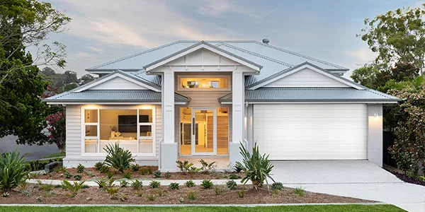 Modern single-story white display home with metal roof, featuring a central entry with warm-lit glass doors and transom window, attached garage on right, landscaped front garden with palm plants, surrounded by mature trees under partly cloudy sky.
