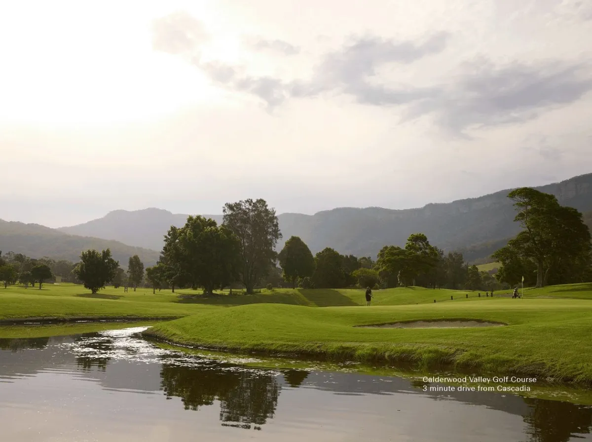 Calderwood Valley Golf Course with lush green fairways, scattered trees, and a stream in the foreground, surrounded by forested mountains under an overcast sky.