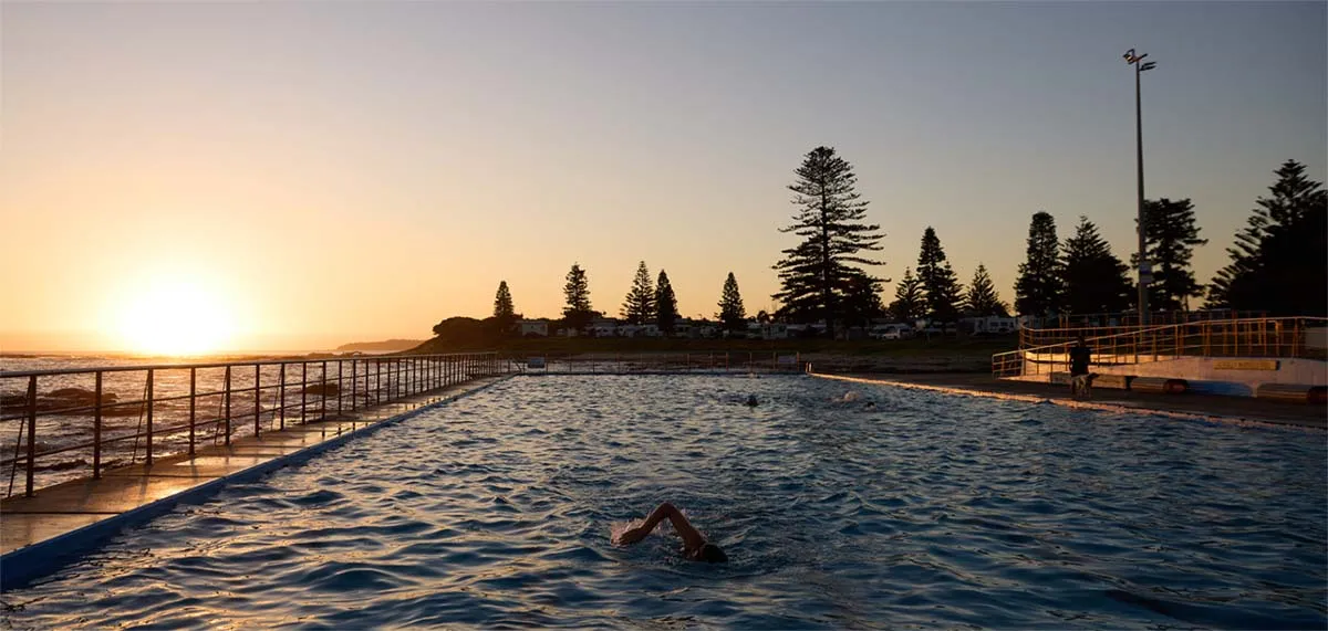 Outdoor oceanside swimming pool at sunrise with swimmers in the water, metal railings along the pool edge, tall pine trees lining the shore, and a street lamp on the right, set against a golden sunrise sky.