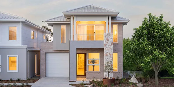 Modern two-story display home with gray brick facade, white garage door, large illuminated windows, metal roof, and manicured landscaping including a green tree on the right side, photographed at dusk in Oran Park.