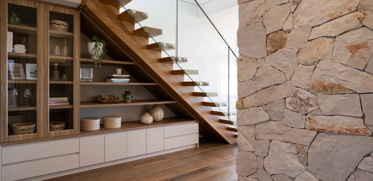 Modern staircase with floating wooden steps and glass railing in a contemporary home interior. Left side features built-in cabinetry with wood and white storage units displaying books and decorative items. Right side shows a textured stone accent wall. Natural light streams in from above, illuminating the warm wood tones and minimalist design.