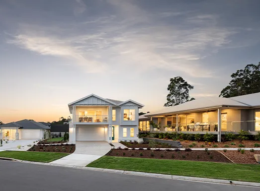Modern two-story white residential home with illuminated windows at dusk, featuring a double garage, manicured landscaping with mulch beds, and adjacent single-story wing, set in a suburban development at twilight.