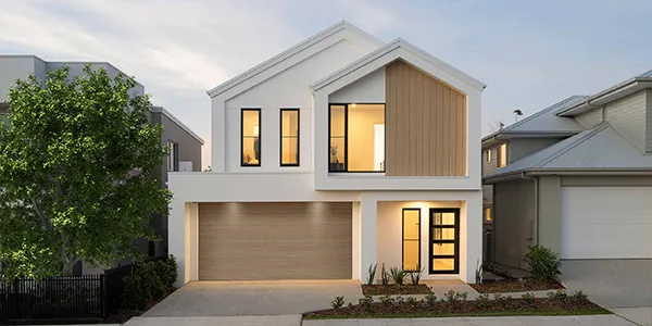 Modern two-story display home with white exterior and timber accent panel, featuring double garage, large front windows with warm lighting, landscaped front garden with green shrubs, located in Oran Park residential estate.