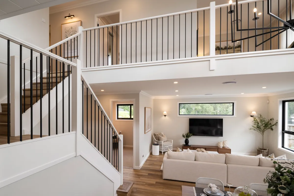 Open-plan living area in Montgomery Homes Waverley display home featuring a two-story space with white and black stair railing, beige sectional sofa, wall-mounted television, wooden flooring, and upper mezzanine level with modern lighting fixtures and neutral décor.