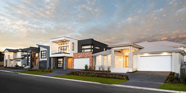 Row of modern display homes in NSW at dusk, featuring contemporary architecture with mixed materials including brick, rendered walls, and dark accents, illuminated by warm interior lighting, positioned along a street with manicured landscaping.