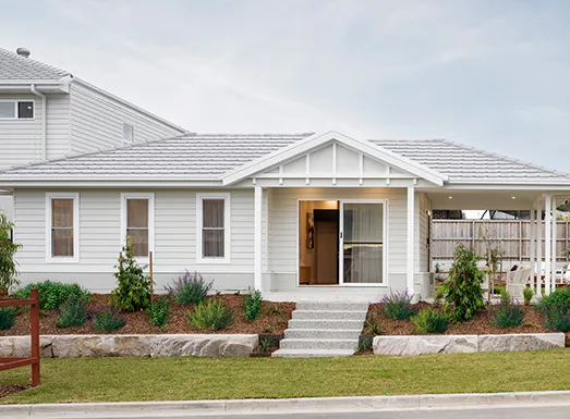 Modern white weatherboard dual-living home with gabled roof, featuring a front entrance with glass doors, manicured garden beds with purple flowering plants, stone edging, and a covered porch area on the right side.