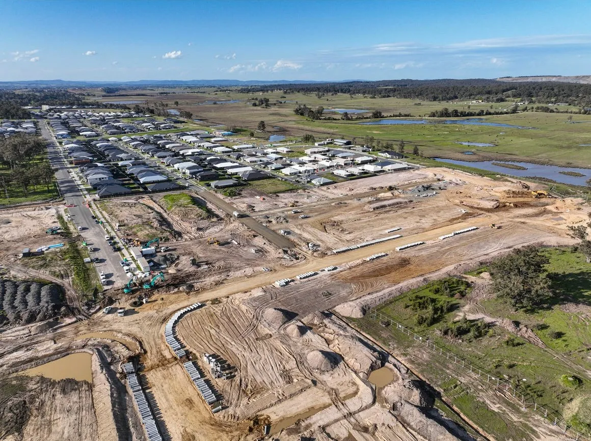 Aerial view of Buchanan Ridge Estate development showing active construction site with earthworks and machinery in foreground, completed residential estates with houses in middle ground, and expansive rural landscape with green fields, trees, and waterways in background under blue sky.