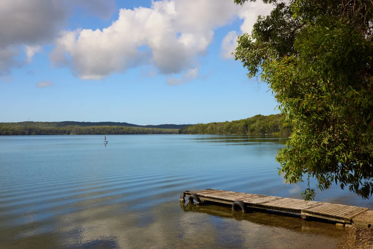 Wooden jetty extending into calm blue water at Crangan Bay Estate on the Central Coast, with forested hills in the background, overhanging green foliage on the right, and a small sailboat visible in the distance under a partly cloudy sky.