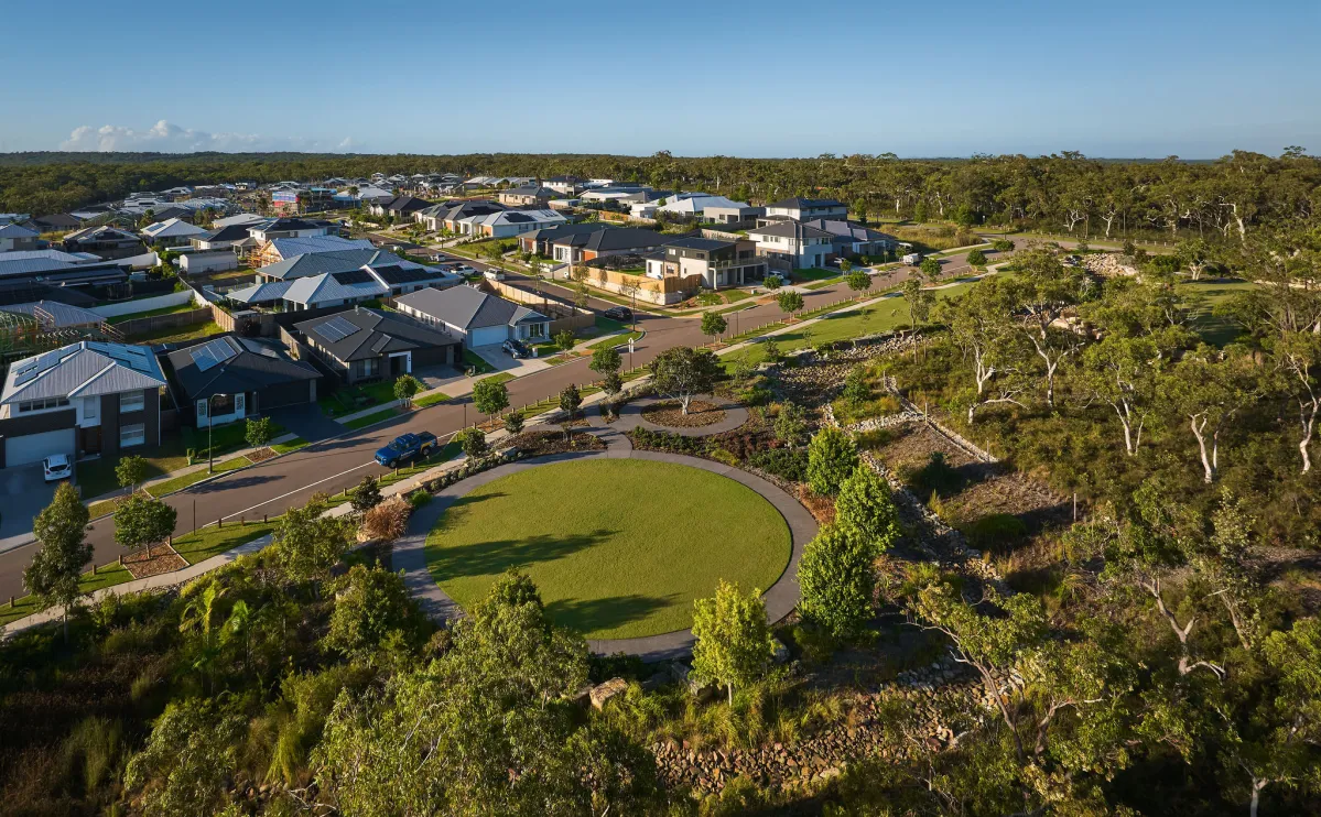 Aerial view of Crangan Bay Estate on Central Coast showing modern residential homes with dark roofs arranged along curved streets, a large circular green space in the foreground, and native eucalyptus trees surrounding the developed area under clear blue sky.