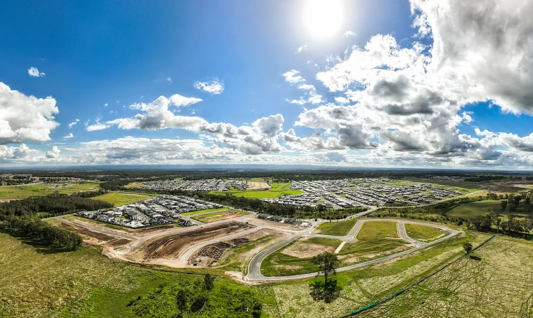 Aerial view of Carmel, Australia showing curved suburban roads through green fields, industrial areas with warehouses, distant town sprawl, and forested areas under a bright blue sky with white clouds.