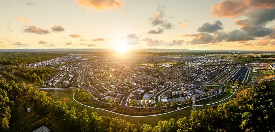 Aerial view of Elara Estate at sunset, showing a sprawling residential development with curved roads, housing subdivisions, and commercial areas surrounded by green bushland and waterways, with the sun setting on the horizon.