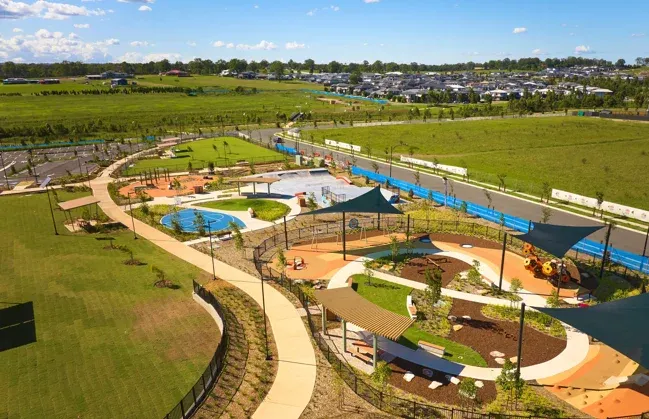 Aerial view of Gables amenity area featuring a modern recreational facility with playgrounds, blue shade structures, winding pathways, landscaped gardens, and sports courts surrounded by expansive green fields and residential development in the background.